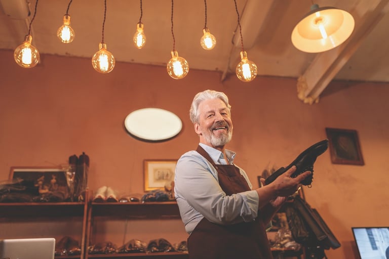 Older man with gray beard working at a workbench in a warm-lit workshop with hanging Edison bulbs overhead