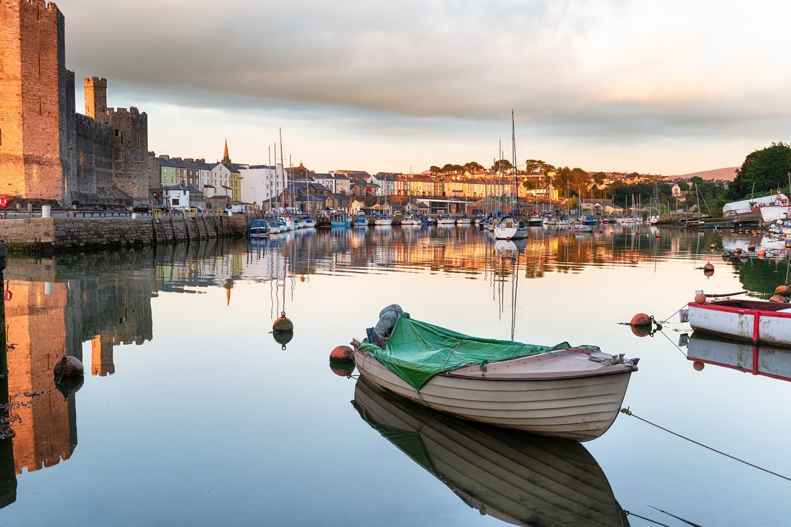 Green and white fishing boats moored in calm harbor water with historic stone buildings and church spire reflected in the background at golden hour