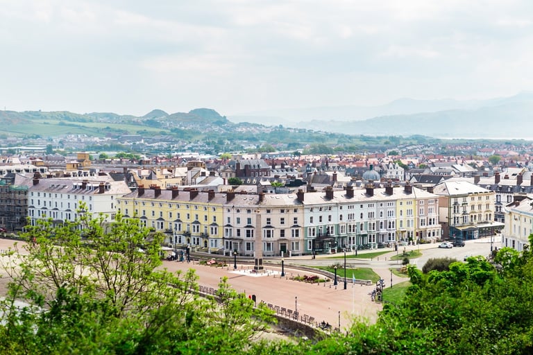 Aerial view of a European city with historic stone buildings, a central plaza, surrounding cityscape, and mountains under a cloudy sky