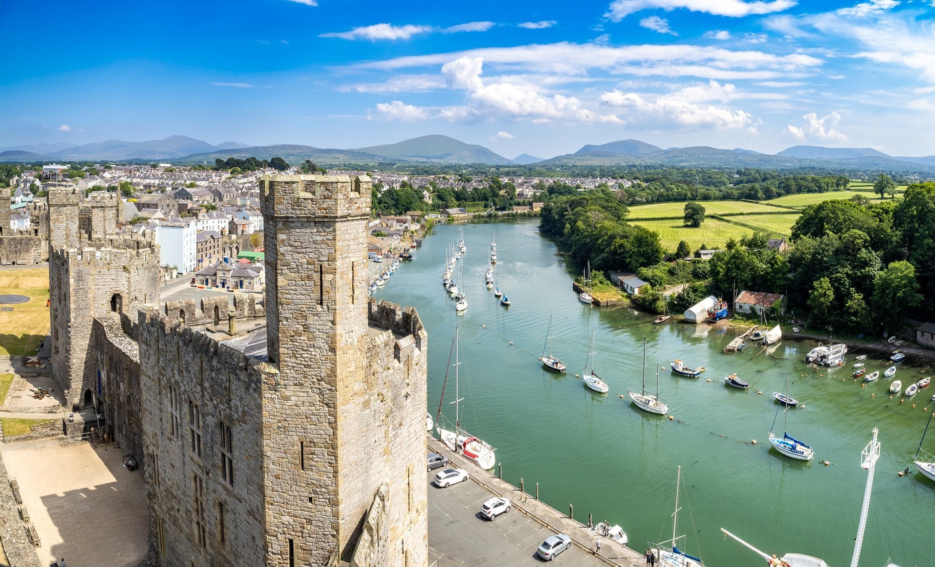 Medieval stone castle walls overlooking a river with moored sailboats, surrounded by green countryside and mountains under blue sky