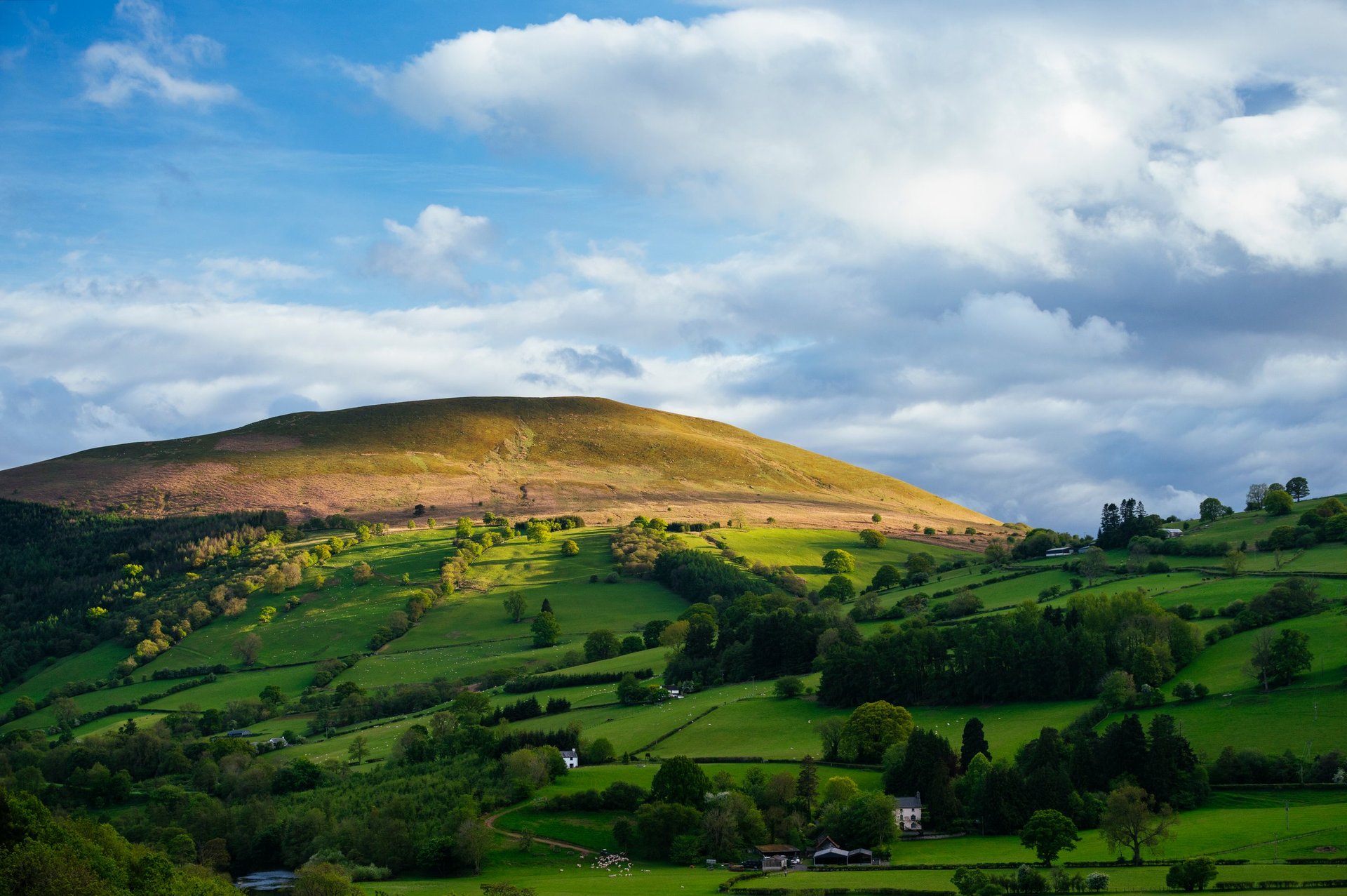Rolling green fields and stone walls beneath a large bare mountain under a partly cloudy sky