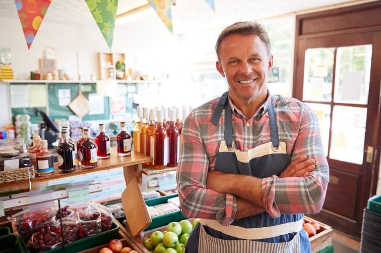 Smiling man in plaid shirt and apron stands in artisanal food shop with jars of preserves and fresh produce displayed