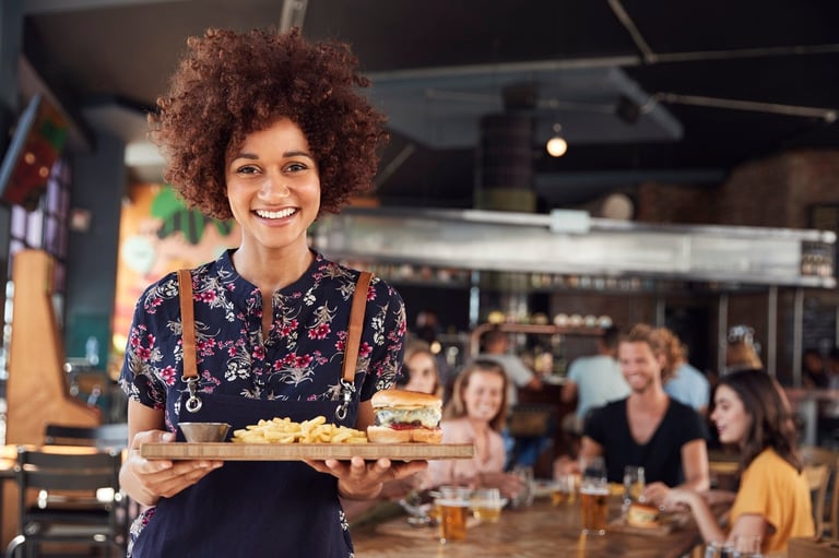 Smiling server with curly hair holding food plate in busy restaurant kitchen with customers in background