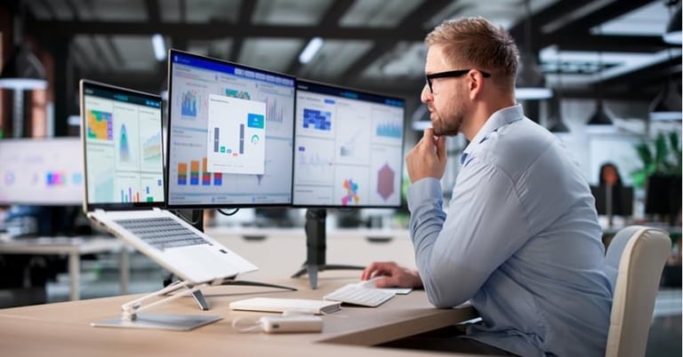 Man in glasses analyzing data on multiple computer monitors in a modern office with charts and analytics displayed