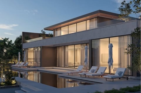 Modern two-story house with glass walls, illuminated interior, infinity pool, and lounge chairs at dusk