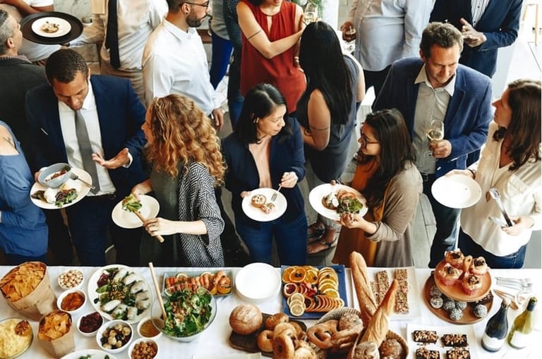Overhead view of a diverse group of people gathered around a table with various foods and plates during a social gathering or event.