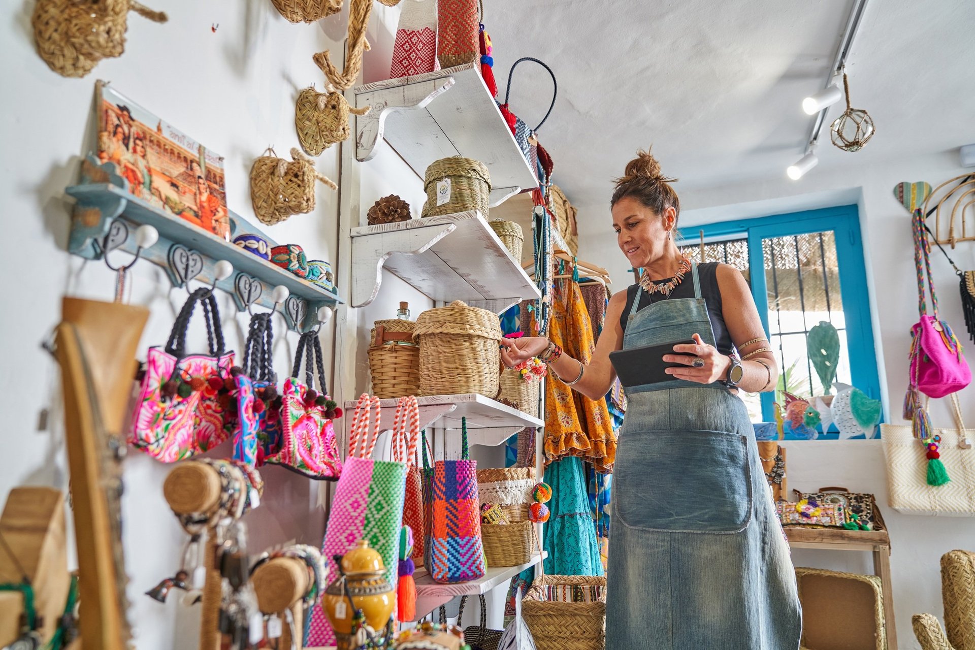 Woman in apron checking inventory at handmade craft shop with hanging baskets, bags, and colorful items on shelves