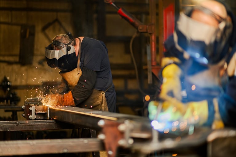 Welder in protective gear and helmet working at an industrial workbench, creating sparks while welding metal.