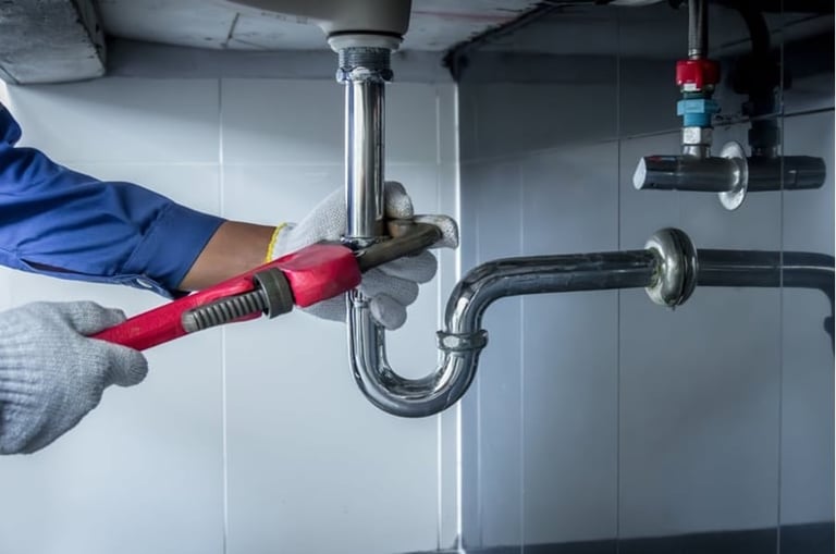 Plumber in blue shirt using a red wrench to tighten pipe connections under a sink with P-trap plumbing